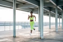 Afroamericana deportista adulta en vibrante ropa deportiva verde enfocándose y corriendo sola a lo largo del paseo marítimo entre columnas metálicas bajo techo - foto de stock