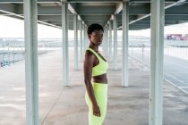 Afrikanisch-amerikanische erwachsene Sportlerin in leuchtend grüner Aktivkleidung, die sich allein am Wasser zwischen Metallsäulen unter dem Dach konzentriert — Stockfoto