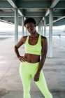 Взрослая афроамериканская спортсменка в ярко-зеленой спортивной одежде, стоящая в одиночестве вдоль набережной среди металлических колонн под крышей — стоковое фото