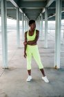 Femme de sport adulte afro-américaine en vêtements de sport vert vif se concentrant debout seul le long du front de mer parmi les colonnes métalliques sous le toit — Photo de stock
