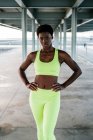 Afrikanisch-amerikanische erwachsene Sportlerin in leuchtend grüner Aktivkleidung, die sich allein am Wasser zwischen Metallsäulen unter dem Dach konzentriert — Stockfoto
