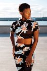 African American woman in flowered tracksuit looking away and thinking while standing with hands on hips alone in sun rays against blurred waterfront — Stock Photo