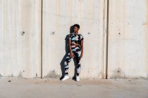 Afrikanisch-amerikanische Sportlerin in geblümter Sportkleidung und weißen Turnschuhen schaut interessiert weg und lacht, während sie nach dem Training in der Stadt allein an einer Betonwand lehnt und sich ausruht — Stockfoto