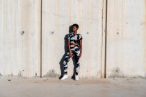 African American female athlete in flowered sport clothes and white sneakers looking away with interest and laughing while standing alone leaning on concrete wall in sunbeams and resting after training in city — Stock Photo