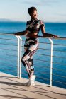 Mujer afroamericana en ropa deportiva con flores y zapatillas blancas mirando hacia otro lado con interés y disfrutando de la vida mientras se apoya en la valla en rayos de sol contra el agua de mar azul calma - foto de stock