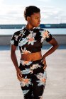 Mujer afroamericana en chándal con flores mirando hacia otro lado y pensando mientras está de pie con las manos en las caderas sola en los rayos del sol contra el borroso paseo marítimo - foto de stock