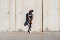 African American female athlete in flowered sport clothes and white sneakers looking away with interest and laughing while standing alone leaning on concrete wall in sunbeams and resting after training in city — Stock Photo
