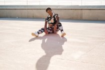 Atleta afroamericana con ropa deportiva florecida y zapatillas blancas mirando a la cámara mientras está sentada sola en la pared de hormigón en rayos de sol y descansando después de entrenar en la ciudad - foto de stock