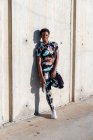 Afrikanisch-amerikanische Sportlerin in geblümter Sportkleidung und weißen Turnschuhen blickt interessiert in die Kamera und lacht, während sie nach dem Training in der Stadt allein an einer Betonwand lehnt und sich ausruht — Stockfoto