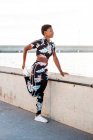 Mujer adulta afroamericana en ropa deportiva con flores que estira los músculos de las piernas mientras está sola y se calienta antes del entrenamiento entre el entorno urbano en un día soleado - foto de stock