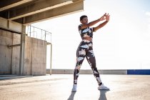 Mulher adulta afro-americana em roupas esportivas floridas alongando os músculos da mão enquanto está sozinha e se aquecendo antes do treinamento entre o ambiente urbano em dia ensolarado — Fotografia de Stock