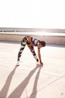 Afrikanisch-amerikanische erwachsene Frau in geblümter Sportbekleidung, die die Beinmuskeln dehnt, während sie allein steht und sich vor dem Training in der städtischen Umgebung bei sonnigem Tag aufwärmt — Stockfoto