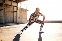 Взрослая афроамериканка в цветущей спортивной одежде растягивает мышцы ног, стоя в одиночестве и разогреваясь перед тренировкой среди городской среды в солнечный день — стоковое фото