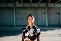 Афроамериканська спортсменка в квітковому спортивному лаві з закритими очима, стоячи на вулиці в сонячних променях до бетонної стіни в місті. — стокове фото