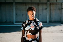 African American female athlete in flowered sportswear looking at camera with challenge while standing alone on street in sunbeams against concrete wall in city — Stock Photo