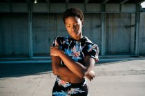 Atleta afro-americana em roupas esportivas floridas com olhos fechados, enquanto está sozinha na rua em raios de sol contra a parede de concreto na cidade — Fotografia de Stock