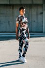 Athlète afro-américaine en vêtements de sport fleuris regardant la caméra avec défi tout en restant seule dans la rue dans les rayons de soleil contre un mur de béton en ville — Photo de stock