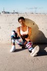 Afrikanisch-amerikanische Athletin in geblümter Sportkleidung und weißen Turnschuhen blickt in die Kamera, während sie allein auf einer Betonwand in Sonnenstrahlen sitzt und sich nach dem Training in der Stadt ausruht — Stockfoto