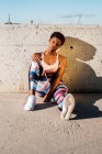 Athlète afro-américaine en vêtements de sport fleuris et baskets blanches regardant la caméra assise seule sur un mur de béton dans les rayons de soleil et se reposant après l'entraînement en ville — Photo de stock