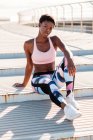 Афроамериканка в цветущей спортивной одежде и белых кроссовках смотрит в камеру, сидя одна на бетонной стене в солнечных лучах и отдыхая после тренировки в городе — стоковое фото