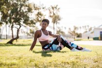 Atleta afroamericana en ropa deportiva colorida y zapatillas blancas mirando hacia otro lado con curiosidad mientras está sentada sobre hierba verde en el césped y descansando después del entrenamiento - foto de stock