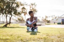 Афро-американская спортсменка в яркой спортивной одежде и белых кроссовках с любопытством смотрит в сторону, сидя на зеленой траве на газоне и отдыхая после тренировки — стоковое фото