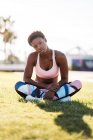 Atleta femenina afroamericana en ropa deportiva colorida y zapatillas blancas mirando a la cámara con curiosidad mientras está sentada sobre hierba verde en el césped y descansando después del entrenamiento - foto de stock