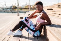 African American young woman in colorful sport clothes and white sneakers looking at camera with curiosity and contemplating while sitting alone on brown wooden stairs on stadium — Stock Photo