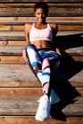 Молодая афроамериканка в разноцветной спортивной одежде и белых кроссовках с любопытством смотрит в камеру и размышляет, сидя в одиночестве на коричневой деревянной лестнице на стадионе — стоковое фото