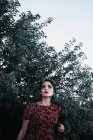 Красива молода жінка дивиться далеко, стоячи під зеленими гілками чагарників на безхмарне небо в сільській місцевості — стокове фото
