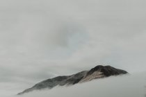 Picos solitários rodeados por nuvens sob o céu cinzento em nevoeiro durante o dia — Fotografia de Stock