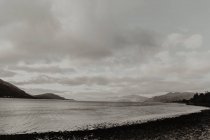 Agua ondulada vacía lavando la costa oscura rodeada de montañas bajo un cielo gris nublado - foto de stock
