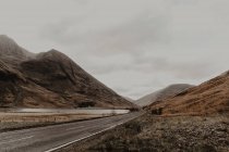 Вузька позначена дорога, що йде вздовж спокійної річки біля підніжжя кам'янистих гір під сірим небом — стокове фото