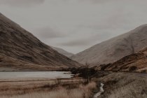 Strada stretta segnata che costeggia il fiume calmo ai piedi delle montagne pietrose sotto il cielo grigio — Foto stock