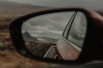 Reflection of car moving on road in front window on empty road along dry hilly valley in gray daytime — Stock Photo