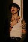 Joven chico elegante con bigote y tatuajes en camiseta blanca apoyada contra la pared, mirando en la cámara - foto de stock