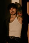 Homem jovem e elegante hipster com tatuagens no topo do tanque branco encostado à parede, olhando na câmera — Fotografia de Stock