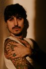 Молодий красивий чоловік з вусами та татуюваннями спирається на стіну в тіні — стокове фото