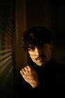 Young melancholic guy in black turtleneck standing next to window with shutters with shadow on face looking in camera — Stock Photo