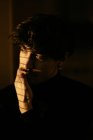 Junger melancholischer Typ in schwarzem Rollkragen, der neben dem Fenster steht, mit Rollläden mit Schatten auf dem Gesicht, der wegschaut — Stockfoto