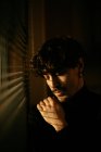 Young melancholic guy in black turtleneck standing next to window with shutters with shadow on face — Stock Photo