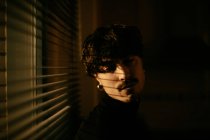 Handsome melancholic guy in black turtleneck standing next to window with shutters, looking down — Stock Photo