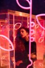 Giovane donna dai capelli lunghi in abito alla moda guardando in macchina fotografica tra i segni al neon in strada città — Foto stock