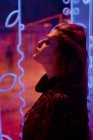 Elegante morena melancólica en luces de letreros de neón apoyados en la pared en la calle de la ciudad - foto de stock
