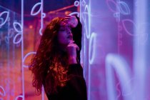 Stylish melancholic brunette in lights of neon signs leaning on wall at city street — Stock Photo