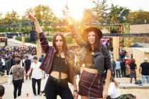 Amigos de moda de cabelos compridos em jaqueta de couro e chapéu dançando levantando as mãos com dois dedos para cima e olhando para a câmera no festival em dia ensolarado — Fotografia de Stock