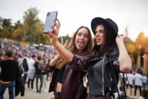 Affascinanti amici allegri in cappello nero che si divertono a sghignazzare e scattare selfie sul telefono cellulare in una giornata luminosa al festival — Foto stock