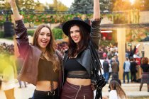 Long haired fashion friends in leather jacket and hat dancing raising hands with two fingers up and looking at camera on festival in sunny day — Stock Photo