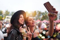 Charming cheerful friends in black hat having fun grimacing and taking selfie on mobile phone in bright day at festival — Stock Photo