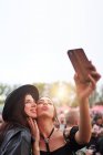 Charming cheerful friends in black hat having fun grimacing and taking selfie on mobile phone in bright day at festival — Stock Photo